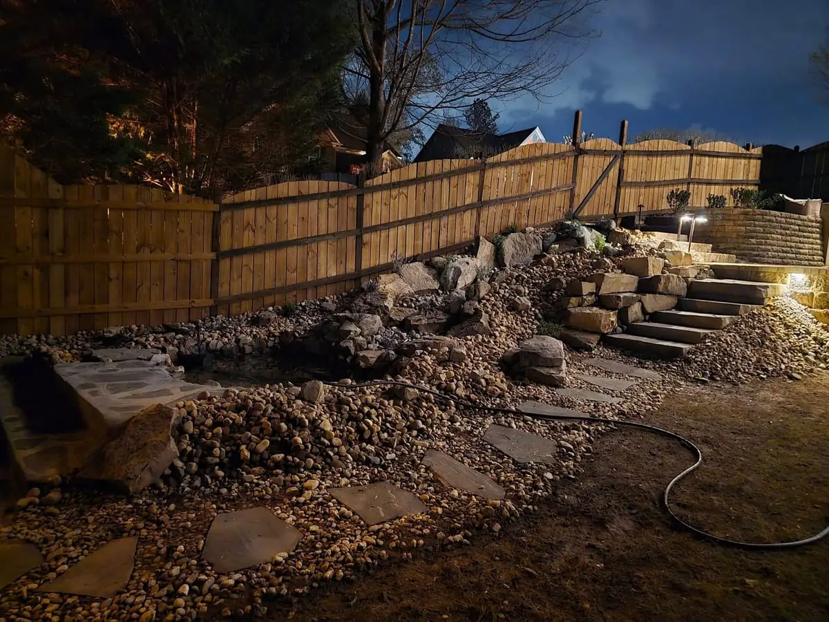Stone steps and boulder retaining wall lit at night along a fenced backyard
