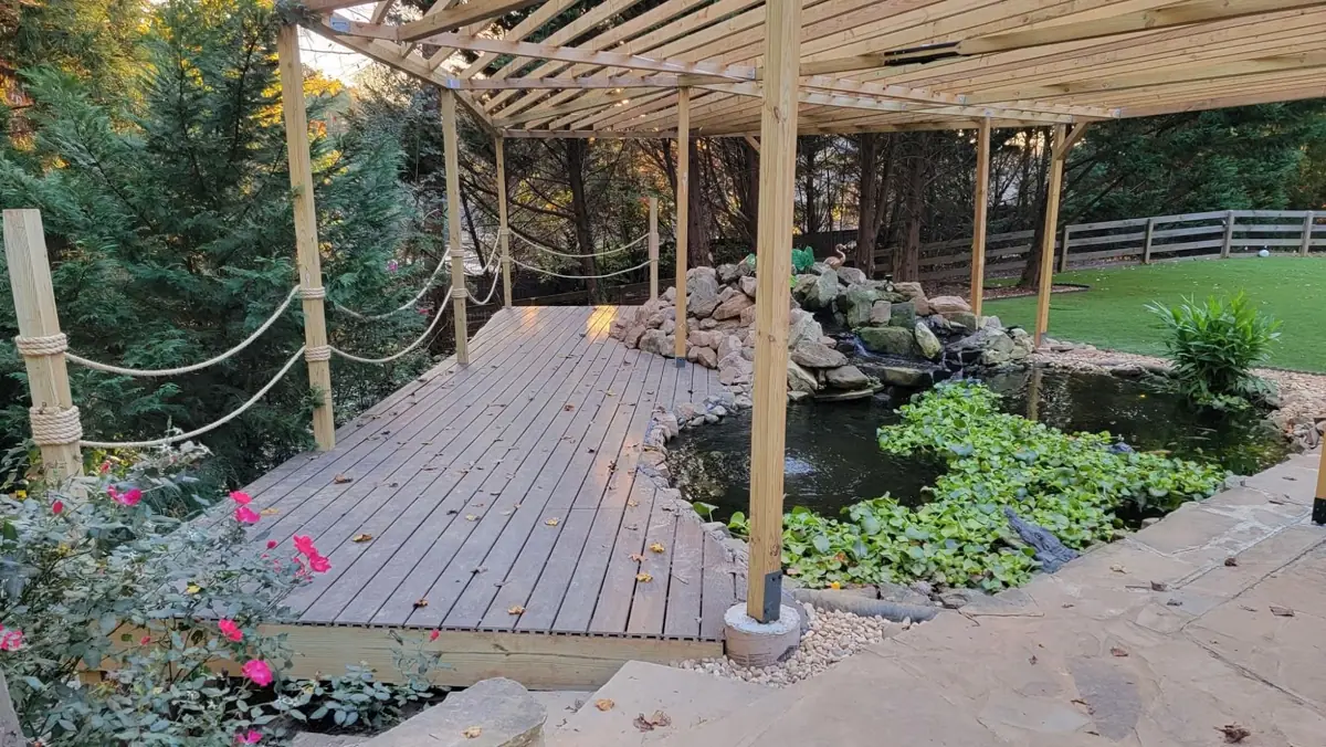 Backyard pond beside a wooden deck under a pergola with rope railings